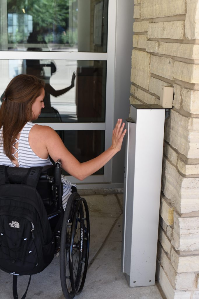 Picture of a young woman in a wheelchair using an automatic door button.