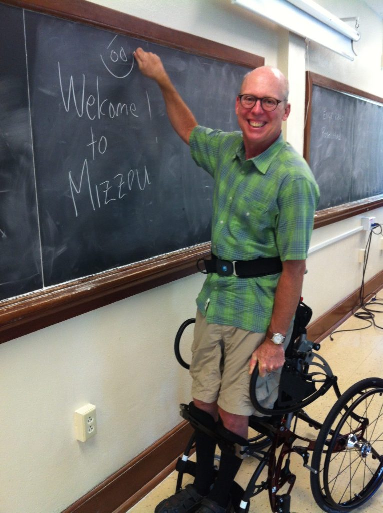 Faculty member using a standing wheelchair, writing on the chalkboard "Welcome to Mizzou"